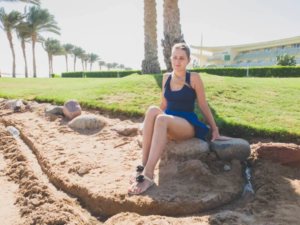 Foto tonificada de una hermosa mujer joven en falda corta sentada en la roca bajo una gran palmera en la playa del mar —  Fotos de Stock