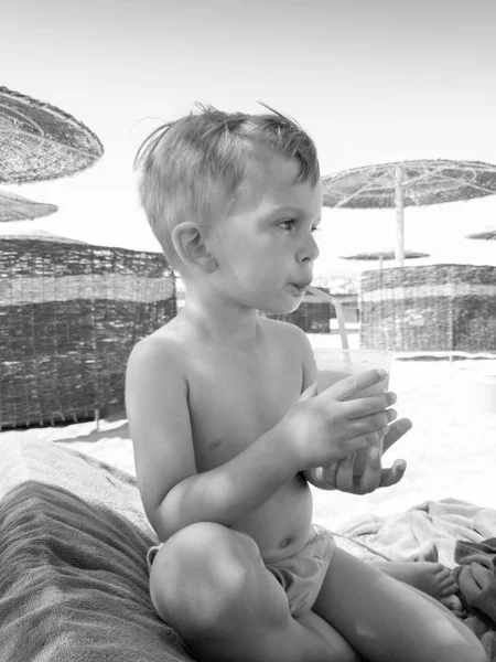 Černý a bílý portrét malého batole, který pije pomerančový džus, když sedí na lenoši u moře. Děti relaxační a mají dobrou dobu během letní dovolené. — Stock fotografie