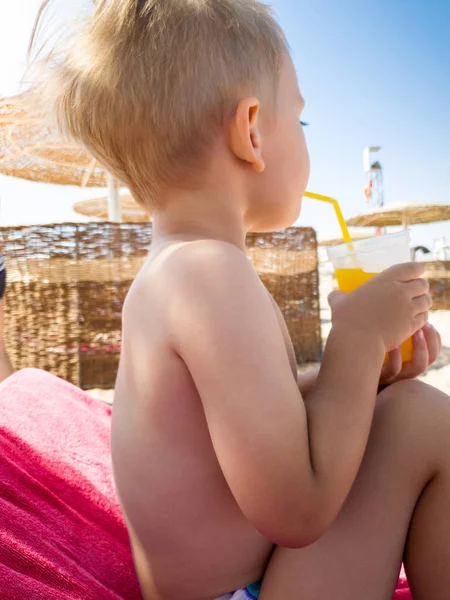 Portrait of little toddler boy sitting on hte sun bed at sea beach and drinking orange juice from straw — Stock Photo, Image