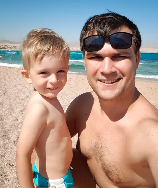 Retrato divertido de padre joven haciendo imagen selfie con su hijo pequeño la playa del mar en soleado brillante. Familia relajante y pasar un buen rato durante las vacaciones de verano . — Foto de Stock