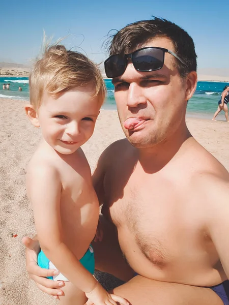 Legrační obraz mladého muže se svým synem, který ukazuje jazyk v kameře, zatímco na mořské pláži dělá fotky. Rodinné relaxace a doba odpočinku během letní dovolené. — Stock fotografie