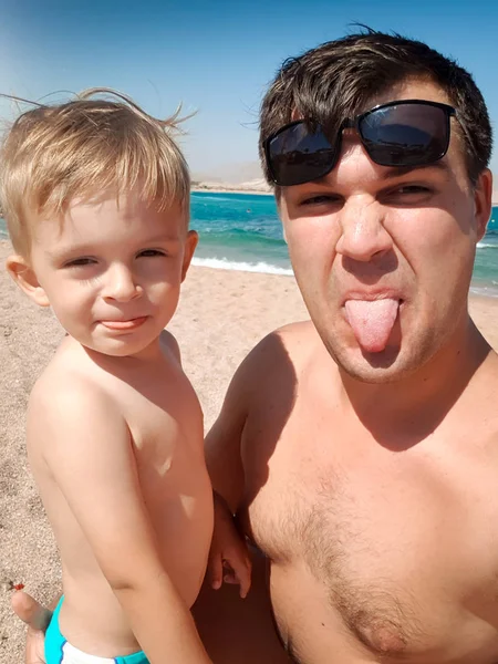 Retrato divertido de padre joven haciendo imagen selfie con su hijo pequeño la playa del mar en soleado brillante. Familia relajante y pasar un buen rato durante las vacaciones de verano . —  Fotos de Stock