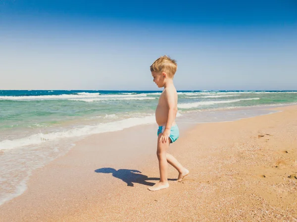 Imagem de adorável menino de 3 anos caminhando na areia da praia quente para o mar. Criança relaxante e se divertindo durante as férias de verão . — Fotografia de Stock