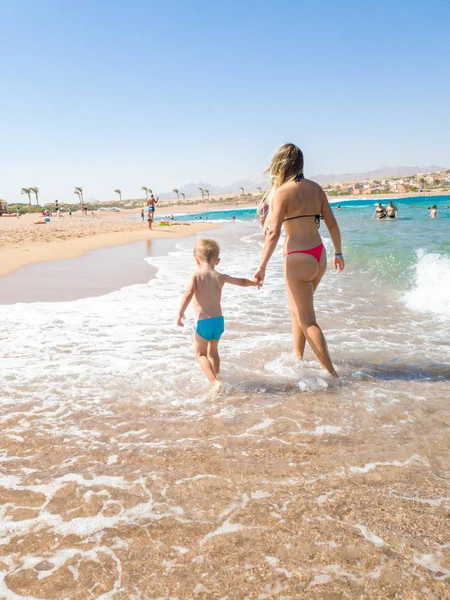 Menino de 3 anos segurando sua jovem mãe à mão e andando na praia do mar. Família relaxante e se divertindo durante as férias de verão . — Fotografia de Stock