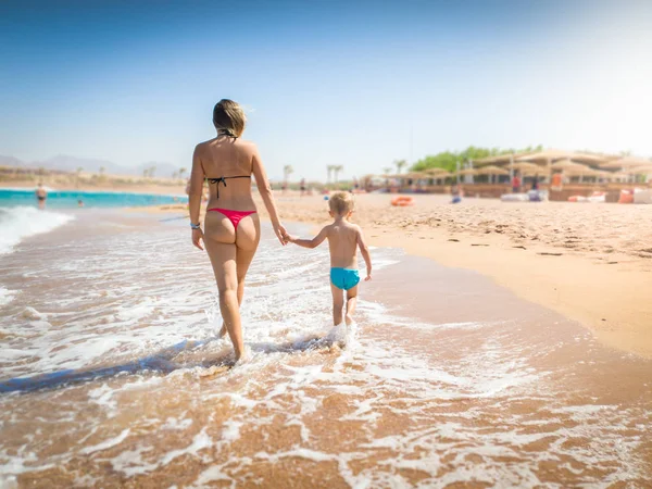 Hermosa madre joven sosteniendo a su pequeño hijo a mano y caminando en la playa de arena de mar en el día soleado brillante. Niño relajante y pasar un buen rato durante las vacaciones de verano . —  Fotos de Stock