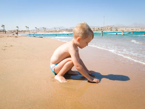 Adorável menino de 3 anos sentado na praia do mar em ondas calmas quentes e brincando com areia molhada. Criança relaxante e se divertindo durante as férias de verão . — Fotografia de Stock