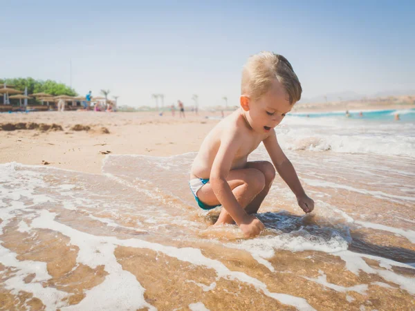 Adorable 3 lat chłopiec Toddler siedzi na plaży morskiej w ciepłych spokojnych fal i grając z mokrym piaskiem. Dziecko relaksujące i dobrze się bawią podczas letnich wakacji. — Zdjęcie stockowe
