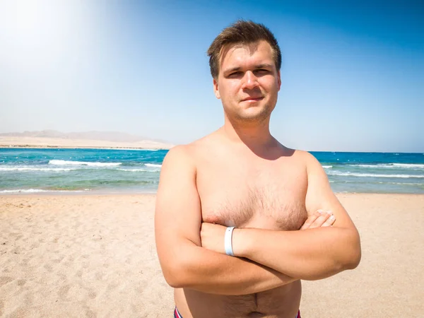 Portræt af smilende muskuløs ung mand poserer på stranden mod hav og blå himmel - Stock-foto