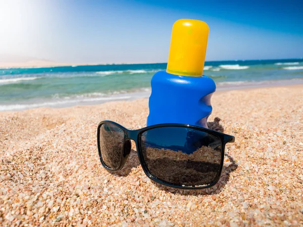 Foto de primer plano de la loción de protección solar UV en botella azul y gafas de sol que se encuentran en la playa de arena en el mar. Imagen perfecta para ilustrar vacaciones de verano . —  Fotos de Stock