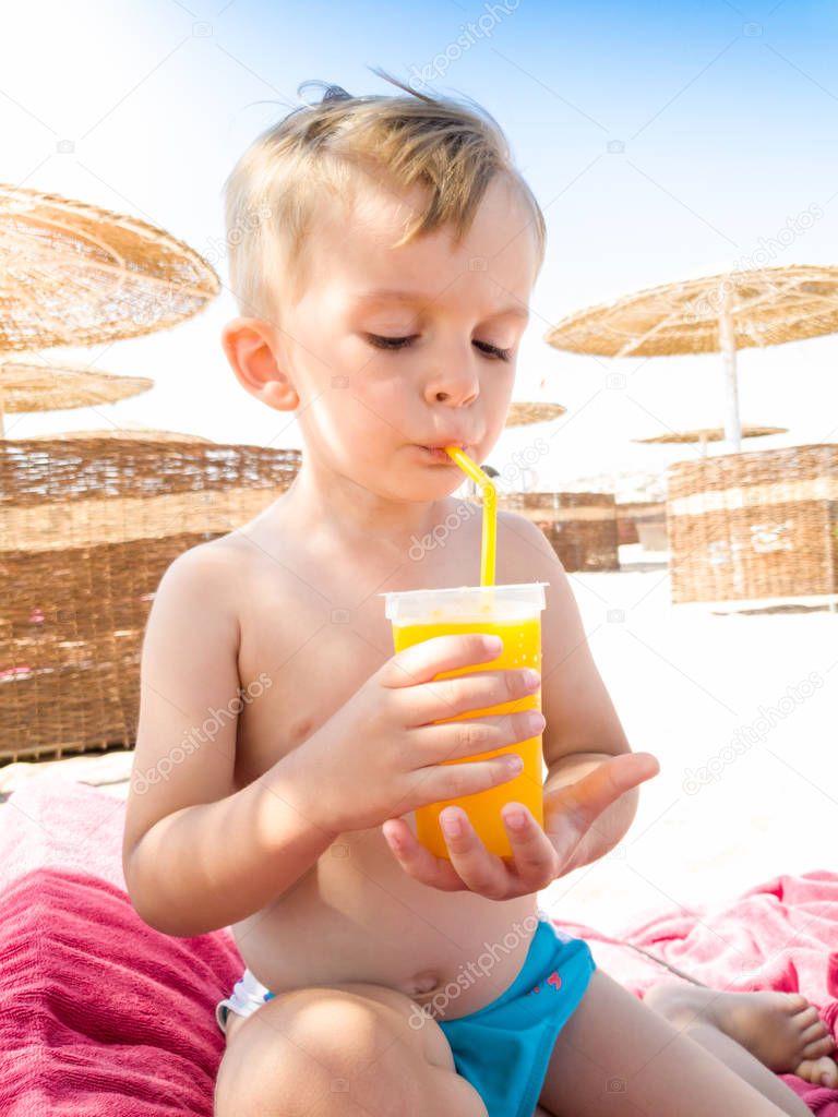 Portrait of little toddler boy sitting on hte sun bed at sea beach and drinking orange juice from straw