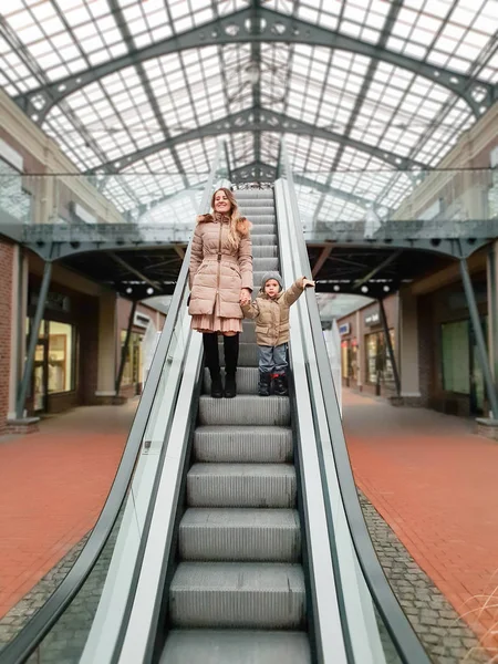 Schöne lächelnde junge Mutter hält ihren kleinen Sohn an der Hand, während sie auf der Rolltreppe im Einkaufszentrum steht — Stockfoto