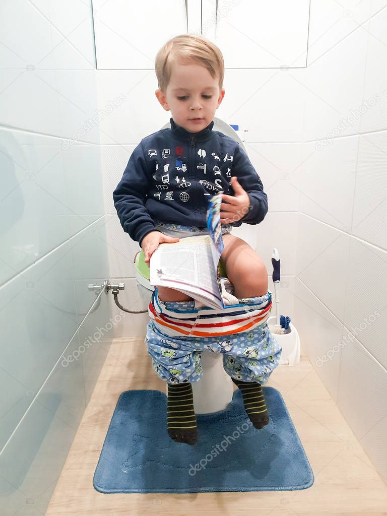 Funny image of little 3 years old toddler boy sitting on the toilet and reading magazine