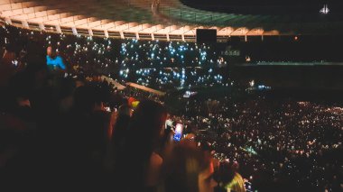 Stadyum koltuklarında oturan ve geceleri rock konseri dinleyen taraftarların odak görüntüsü.