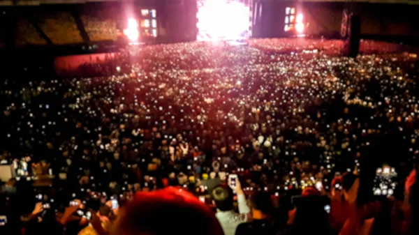 Defocused photo of people listening and watching big rock concert on music festival at big stadium. Crowd of fans sitting on the tribunes at night. Illuminated scene with light beams and lasers — Stock Photo, Image
