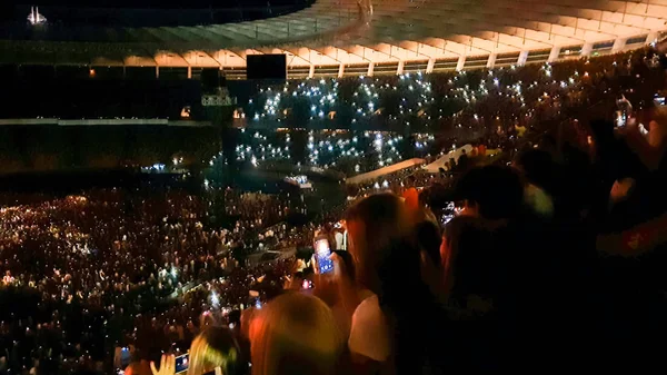 Fotografie lidí, kteří poslouchají a sledují velký Rock koncert na hudebním festivalu na velkém stadionu. Dav fanoušků sedících v noci na tribunách. Osvětlená scéna se světelnými trámy a lasery — Stock fotografie