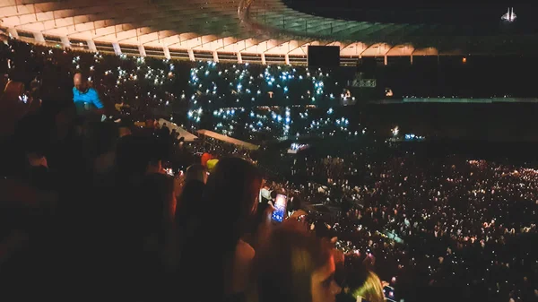 Out of focus image of big crowd of fans sitting on stadium seats watching and listening rock concert at night. — Stock Photo, Image