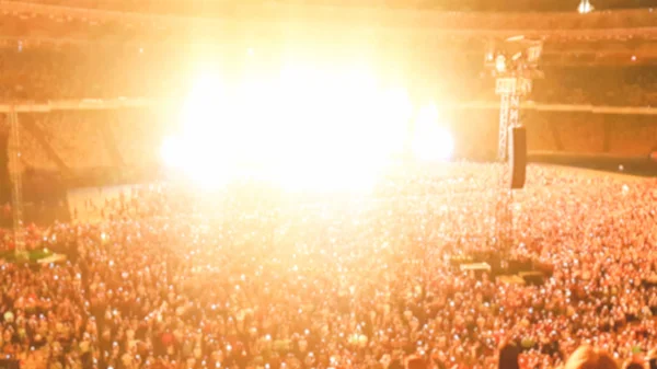 Foto desenfocada de personas escuchando y viendo un gran concierto de rock en un festival de música en un gran estadio. Multitud de fans sentados en las tribunas por la noche. Escena iluminada con rayos de luz y láseres — Foto de Stock