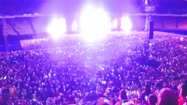 Out of focus image of big crowd of fans sitting on stadium seats watching and listening rock concert at night. — Stock Photo, Image