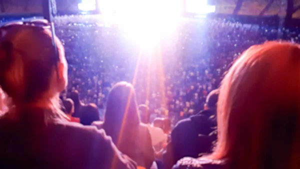 Rear view out of focus photo of people sitting on the music concert at night. Bright colorful light beams illuminating scene. — Stock Photo, Image