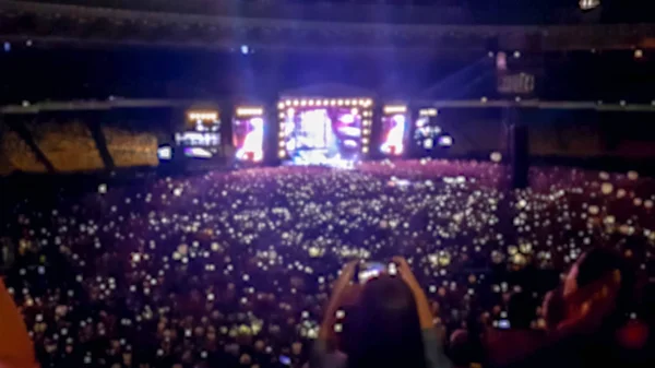 Foto desenfocada de personas escuchando y viendo un gran concierto de rock en un festival de música en un gran estadio. Multitud de fans sentados en las tribunas por la noche. Escena iluminada con rayos de luz y láseres — Foto de Stock