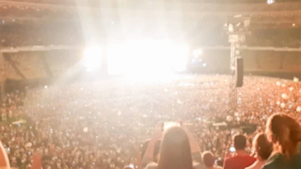Out of focus image of big crowd of fans sitting on stadium seats watching and listening rock concert at night. — Stock Photo, Image