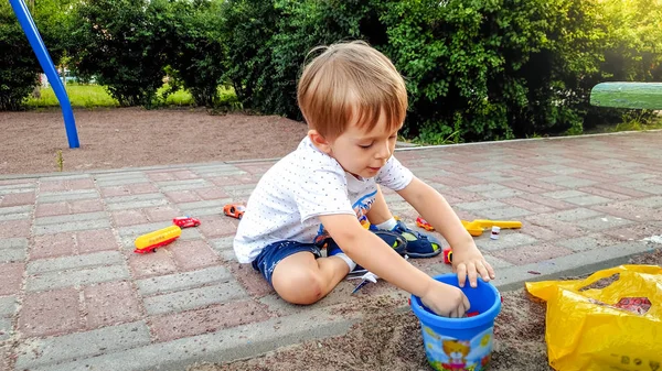 Petit garçon de 3 ans en t-shirt et short assis sur le sol à l'aire de jeux et jouant avec beaucoup de jouets colorés — Photo