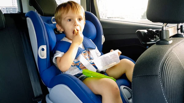 Retrato de un adorable niño sentado en un asiento de seguridad para niños y comiendo galletas — Foto de Stock