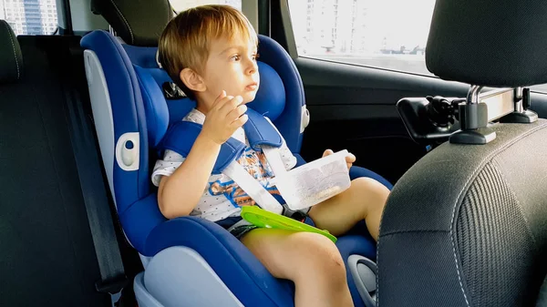 Pequeño niño sintiendo hambre y comiendo mientras viaja en coche en asiento de seguridad para niños —  Fotos de Stock