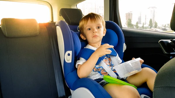 Retrato de un niño de 3 años sentado en el asiento de seguridad infantil en el coche y comiendo galletas. Niños viajando en automóvil —  Fotos de Stock