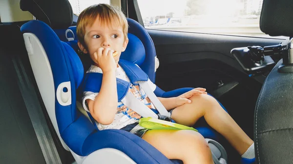 Retrato de un niño de 3 años sentado en el asiento de seguridad infantil en el coche y comiendo galletas. Niños viajando en automóvil — Foto de Stock