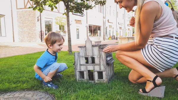 Image tonique d'un petit garçon souriant assis avec une jeune mère dans un parc à regarder à l'intérieur d'une petite maison en bois — Photo