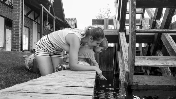 Foto en blanco y negro del niño lindo con la madre arrodillada en el pequeño río tranquilo con molino de agua y mirando la superficie del agua —  Fotos de Stock