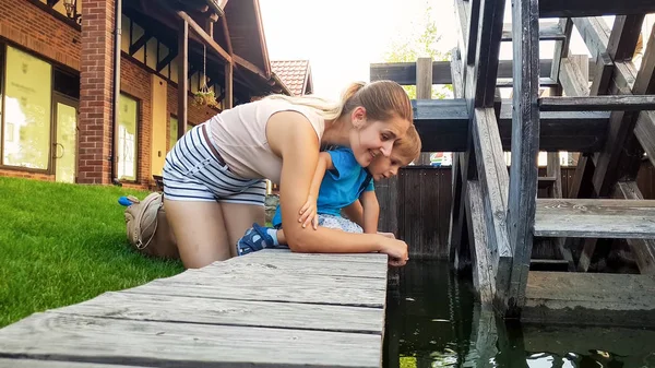 Portrait d'une jeune mère souriante heureuse avec son petit enfant touchant l'eau dans le canal à côté d'un vieux moulin à eau en bois — Photo