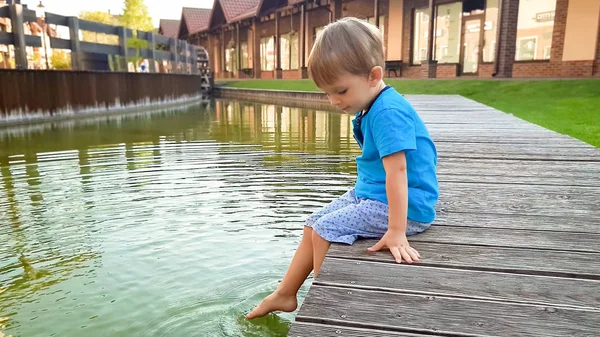 Foto de bonito menino de 3 anos sentado na margem do rio no canal de água na cidade velha e salpicando água com os pés . — Fotografia de Stock