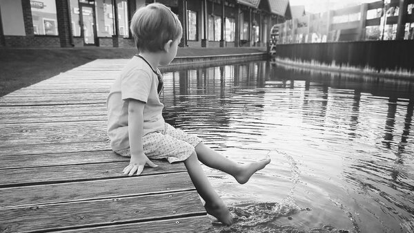 Black and white image of 3 years old toddler boy sitting on the pier at small river in town and holding feet in water