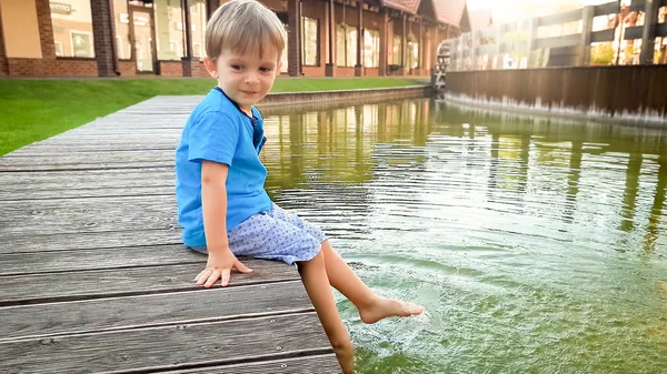 Urocza 3-letnia chłopiec maluch siedzi na brzegu rzeki w małym europejskim mieście w słonecznym letnim dniu — Zdjęcie stockowe