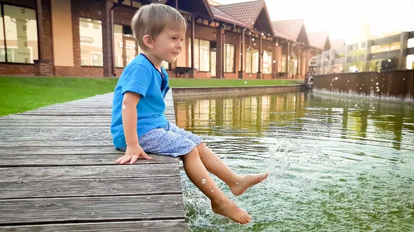 Portret adorable uśmiechnięty chłopiec chłopca siedzi na rzece w małym miasteczku i trzyma stopy w wodzie — Zdjęcie stockowe