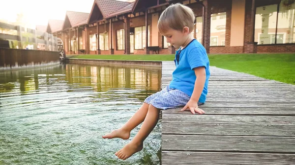 Eski şehirde su kanalında nehir kıyısında oturan sevimli 3 yaşındaki küçük çocuk fotoğraf ve ayakları ile su sıçrayan. — Stok fotoğraf