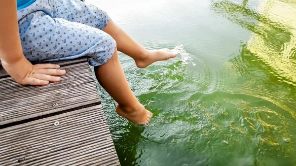 Κοντινό πλάνο του παιδιού που κάθεται στην ξύλινη προβλήτα στο tiver και κρατώντας τα πόδια στο νερό. Παιδιά που παίζουν και πιτσιλίζουν το νερό με τα πόδια — Φωτογραφία Αρχείου
