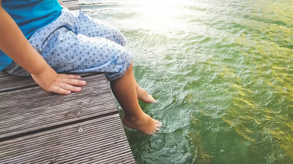 Foto de primer plano de un niño de 3 años sentado en el muelle de madera y sosteniendo sus pies en el agua del río. CHILD salpicaduras en el lago con las piernas — Foto de Stock