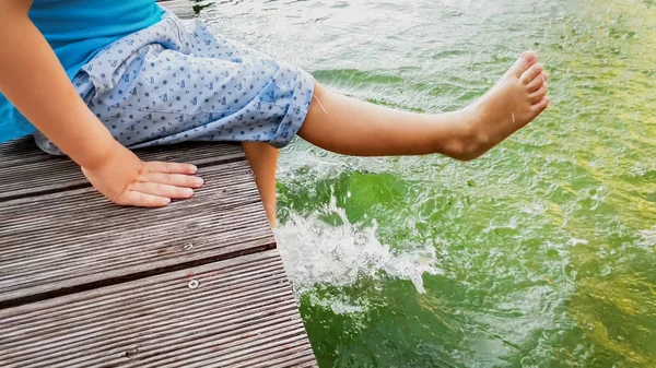 Κοντινό πλάνο του ξυπόλυτου μικρού παιδιού που κάθεται στην ξύλινη γέφυρα στη λίμνη και πιτσιλίζει το νερό με τα πόδια του — Φωτογραφία Αρχείου