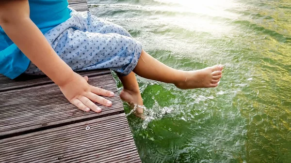 Photo rapprochée de 3 ans petit garçon assis sur la jetée en bois et tenant ses pieds dans l'eau de la rivière. Éclaboussures de poussin dans le lac avec les jambes — Photo
