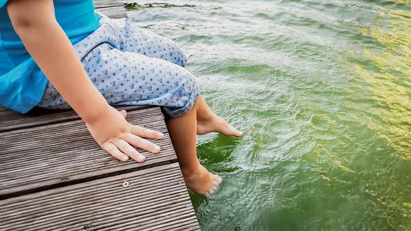 Closeup image of child sitting on the wooden pier at tiver and holding feet in water. Kids playing and splashing water with legs