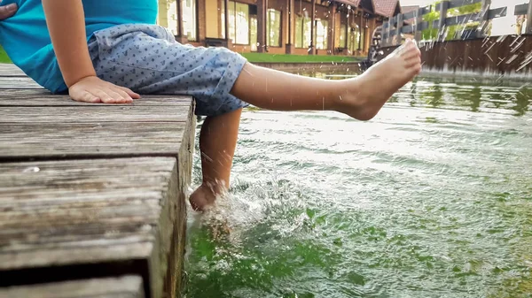 Κοντινό πλάνο φωτογραφίας ενός μικρού αγοριού 3 ετών που καθόταν στην ξύλινη προβλήτα και κρατούσε τα πόδια του στο νερό του ποταμού. Παιδικό πιτσίλισμα σε λίμνη με πόδια — Φωτογραφία Αρχείου