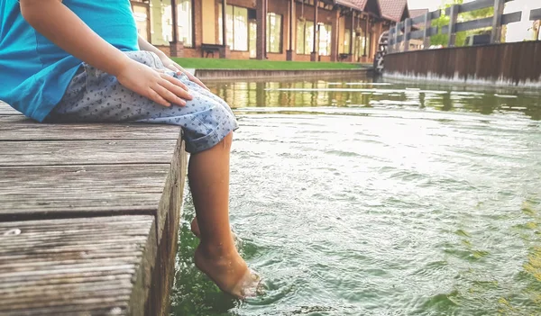 Closeup image of child sitting on the wooden pier at tiver and holding feet in water. Kids playing and splashing water with legs — Stock Photo, Image