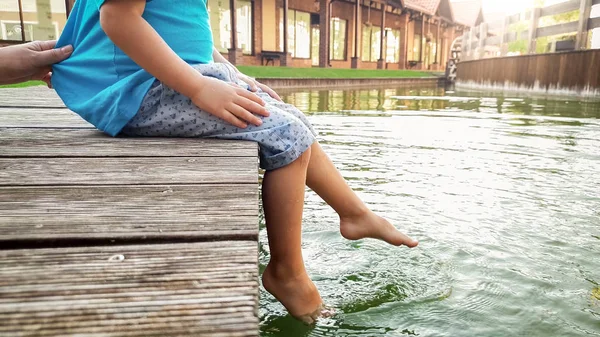 裸足の小さな幼児の少年が湖の木製の橋の上に座って、彼の足で水をはね合うクローズアップ画像 — ストック写真