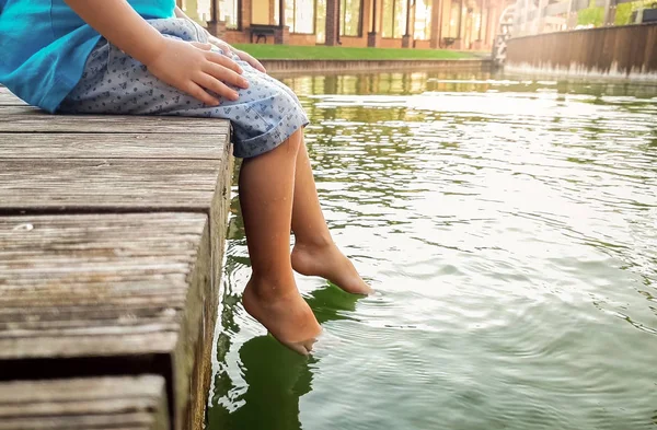 Closeup photo of 3 years old little boy sitting on the wooden pier and holding his feet in river water. CHild splashing in lake with legs — Stock Photo, Image