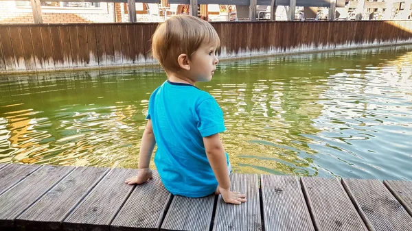 Adorable niño de 3 años sentado en la orilla del río en la pequeña ciudad europea en el soleado día de verano — Foto de Stock