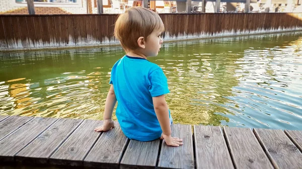 Sevimli gülümseyen yürümeye başlayan küçük çocuk portresi küçük bir kasabada nehirde oturan ve suda ayakları tutarak — Stok fotoğraf