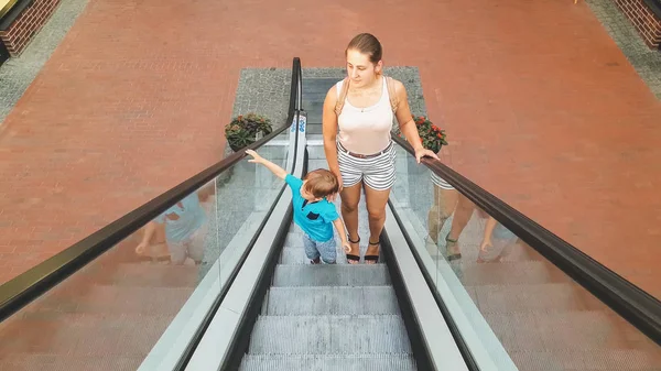 Alışveriş merkezinde yürüyen merdiven üzerinde duran genç anne ile sevimli yürümeye başlayan çocuk — Stok fotoğraf